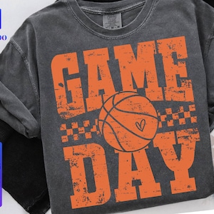 Puede incluir: Camiseta gris oscuro con las palabras "GAME DAY" en letras naranjas desgastadas, con una pelota de baloncesto naranja con un coraz&oacute;n. La camiseta tambi&eacute;n tiene un peque&ntilde;o patr&oacute;n de cuadros.