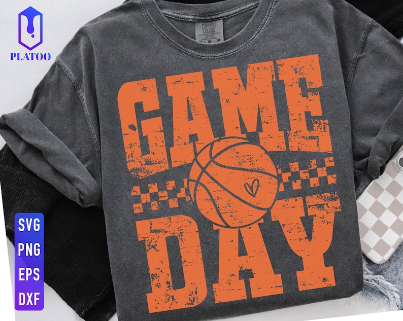 Puede incluir: Camiseta gris oscuro con las palabras "GAME DAY" en letras naranjas desgastadas, con una pelota de baloncesto naranja con un coraz&oacute;n. La camiseta tambi&eacute;n tiene un peque&ntilde;o patr&oacute;n de cuadros.