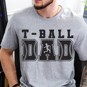 Puede incluir: Una camiseta gris con el texto "T-BALL DAD" en fuente negra desgastada. La palabra "DAD" presenta un dise&ntilde;o de b&eacute;isbol con una silueta de bateador. La camiseta es de estilo casual.