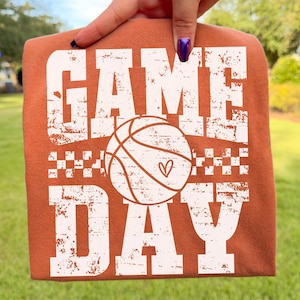 Puede incluir: Una camiseta naranja quemado con las palabras "GAME DAY" en letras blancas desgastadas. Una pelota de baloncesto con un coraz&oacute;n est&aacute; centrada entre las palabras. La camiseta est&aacute; sujeta por una mano, con un fondo verde.