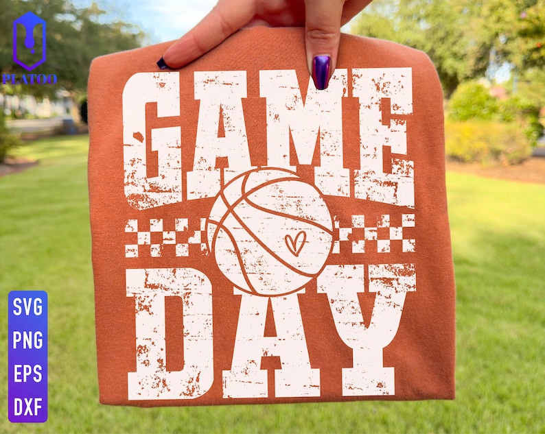 Puede incluir: Una camiseta naranja quemado con las palabras "GAME DAY" en letras blancas desgastadas. Una pelota de baloncesto con un coraz&oacute;n est&aacute; centrada entre las palabras. La camiseta est&aacute; sujeta por una mano, con un fondo verde.