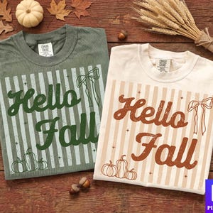 Op de afbeelding: Twee T-shirts met ronde hals, één saliegroen en één crème, elk met een verticaal gestreepte achtergrond, een strikillustratie, drie pompoenomtrekken en de tekst "Hello Fall" in een verweerd lettertype met kleine steraccenten.