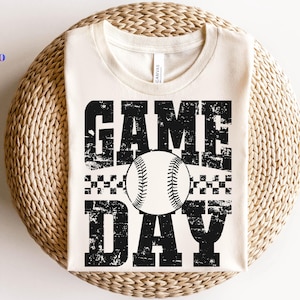 May include: A cream-coloured t-shirt with the words "GAME DAY" in distressed black lettering, featuring a baseball graphic. The shirt is displayed on a woven, circular surface. The shirt is a baseball-themed item.