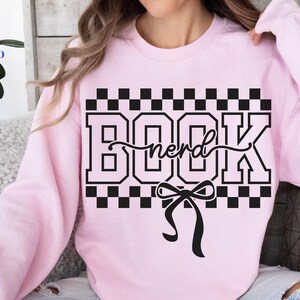 May include: A light pink sweatshirt with a black and white checkered design and the words "BOOK nerd" in a bold font. A black bow is below the text. The sweatshirt is a casual style.