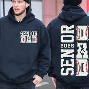 Puede incluir: Una sudadera con capucha azul oscuro con el texto "Senior Dad 2026" en letras blancas. El texto está rodeado de gráficos de temática de béisbol, incluyendo una pelota de béisbol, un bate de béisbol y un jugador de béisbol.