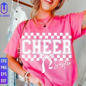 Op de afbeelding: Een roze t-shirt met de woorden "CHEER Sister" in een wit blokletterontwerp. Het ontwerp bevat een dambordpatroon en een strik. Het shirt heeft korte mouwen en lijkt gemaakt van een zacht materiaal.