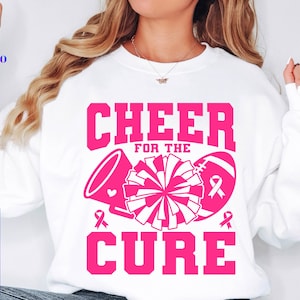 May include: White sweatshirt with the words "CHEER FOR THE CURE" in pink, along with a megaphone, pom-pom, and football. The design includes pink ribbons, symbolizing breast cancer awareness. The sweatshirt is a crew neck.