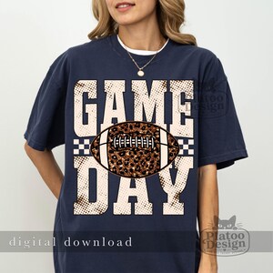 May include: A navy blue short-sleeve t-shirt featuring a large graphic print with the text GAME DAY in a distressed beige font. Centered between the words is a football illustration with a brown leopard print pattern and white laces.