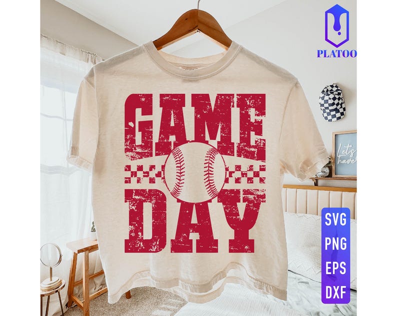 May include: A cream-coloured t-shirt featuring the words "GAME DAY" in distressed red lettering and a baseball graphic. The shirt is hanging on a wooden hanger. The design includes a baseball and a checkered pattern.