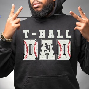 Puede incluir: Sudadera con capucha negra con el texto "T-BALL DAD" en letras blancas. La palabra "DAD" est&aacute; dise&ntilde;ada como una pelota de b&eacute;isbol con costuras rojas. Una silueta de un jugador de b&eacute;isbol est&aacute; en el centro de la palabra "DAD".