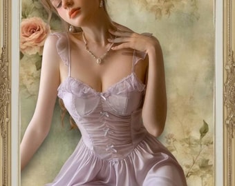 May include: A woman wearing a light purple satin slip dress with lace trim and a bow detail at the bust. The dress has a fitted bodice and a flowing skirt. The woman is sitting in a chair with a floral background.