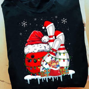 May include: Black t-shirt featuring a Christmas-themed bowling design. A red bowling ball wears a Santa hat and is wrapped in Christmas lights. Bowling pins are also wearing Santa hats and are decorated with lights. Presents and snowflakes complete the festive scene.