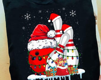 May include: Black t-shirt with a Christmas bowling design. A red bowling ball wearing a Santa hat is wrapped in Christmas lights. Bowling pins also wear Santa hats and are decorated with lights. Presents and snowflakes complete the festive scene.