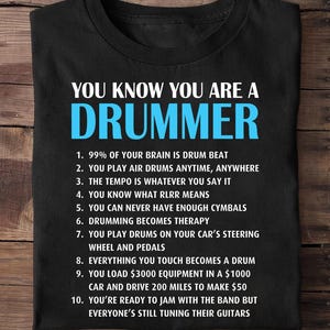 May include: Black t-shirt with white and blue text: "YOU KNOW YOU ARE A DRUMMER". The shirt lists ten humorous statements about drummers, including playing air drums and loading expensive equipment.