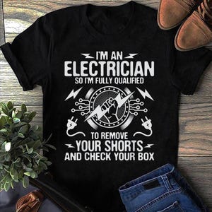 Op de afbeelding: Een zwart t-shirt met korte mouwen en witte tekst die luidt I'M AN ELECTRICIAN SO I'M FULLY QUALIFIED TO REMOVE YOUR SHORTS AND CHECK YOUR BOX, vergezeld van een afbeelding van een vuist die een bliksemschicht en stekkers vasthoudt.