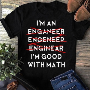 May include: Black t-shirt with white text: "I'M AN ENGINEER I'M GOOD WITH MATH." The word "engineer" is crossed out multiple times with red lines, correcting the spelling. The shirt is on a wooden surface with jeans and shoes.