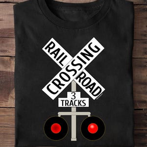 May include: Black t-shirt with a white railroad crossing sign that reads "RAIL CROSSING ROAD". Below the sign is a rectangle with "3 TRACKS". Two red circles are below the rectangle.