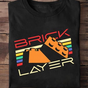 Pode incluir: Uma t-shirt preta de gola redonda com uma impressão gráfica com o texto BRICK LAYER em vermelho e creme. O design inclui uma ilustração de um bloco de construção de plástico laranja sobre um fundo de riscas horizontais vermelhas, amarelas, verdes e azuis.