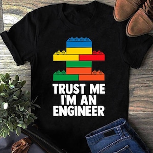May include: A black short-sleeved t-shirt featuring a graphic of colorful interlocking plastic building blocks stacked in a tower. Below the blocks, the white text reads: TRUST ME I'M AN ENGINEER. The design uses blue, yellow, red, green, and orange colors.