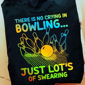 May include: Black t-shirt with a rainbow graphic of bowling pins and a bowling ball. The text "THERE IS NO CRYING IN BOWLING... JUST LOT'S OF SWEARING" is printed on the shirt.
