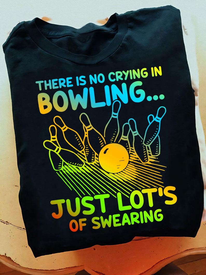 May include: Black t-shirt with a rainbow graphic of bowling pins and a bowling ball. The text "THERE IS NO CRYING IN BOWLING... JUST LOT'S OF SWEARING" is printed on the shirt.