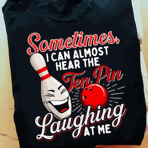 May include: Black t-shirt featuring a bowling pin with a smiling face, a red bowling ball, and the text "Sometimes, I can almost hear the Ten Pin Laughing at Me" in red and white. The design is centered on the shirt.