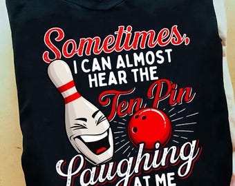 May include: Black t-shirt featuring a bowling pin with a smiling face, a red bowling ball, and the text "Sometimes, I can almost hear the Ten Pin Laughing at Me" in red and white. The design is centered on the shirt.