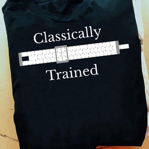 Könnte beinhalten: Gefaltetes schwarzes T-Shirt mit dem weißen Schriftzug "Classically Trained" und einer weißen Rechenschieber-Grafik mit grauen Akzenten. Das Design ist auf der Vorderseite des T-Shirts aufgedruckt.