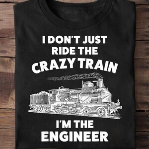 May include: Black t-shirt with the text "I DON'T JUST RIDE THE CRAZY TRAIN I'M THE ENGINEER" in white. A detailed black and white illustration of a steam train is below the text. The shirt is folded on a wooden surface.