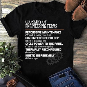 May include: Black t-shirt featuring the text "Glossary of Engineering Terms" with amusing definitions for engineering concepts. The shirt includes a gear graphic and is shown with blue jeans and brown shoes.