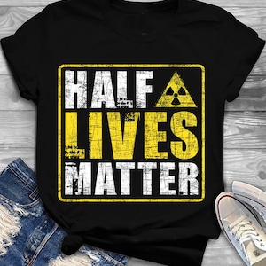 Könnte beinhalten: Schwarzes T-Shirt mit einem verblichenen gelb-weißen Aufdruck. Der Aufdruck lautet "HALF LIVES MATTER" mit einem Strahlungssymbol. Das Shirt wird mit blauen Jeans und weißen Turnschuhen kombiniert.