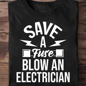 Puede incluir: Camiseta negra con texto blanco: "SAVE A FUSE BLOW AN ELECTRICIAN". El texto está dispuesto en tres líneas con gráficos de rayos. La camiseta está doblada sobre una superficie de madera.
