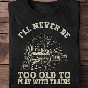 May include: Black t-shirt featuring a vintage train illustration with the text "I'll Never Be Too Old To Play With Trains" in a distressed font. The design includes a detailed steam engine and radiating lines.
