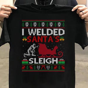 Op de afbeelding: Zwart T-shirt met een kerstontwerp. Het shirt toont de tekst "I Welded Santa's Sleigh" in wit en rood, samen met een afbeelding van een lasser, de slee van de kerstman en feestelijke randen in groen en rood.