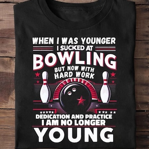May include: Black t-shirt with a bowling-themed graphic. The design includes bowling pins, a bowling ball, and text that reads "WHEN I WAS YOUNGER I SUCKED AT BOWLING BUT NOW WITH HARD WORK DEDICATION AND PRACTICE I AM NO LONGER YOUNG."