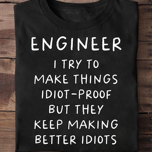 Peut inclure: T-shirt noir avec texte blanc : "ENGINEER I TRY TO MAKE THINGS IDIOT-PROOF BUT THEY KEEP MAKING BETTER IDIOTS."