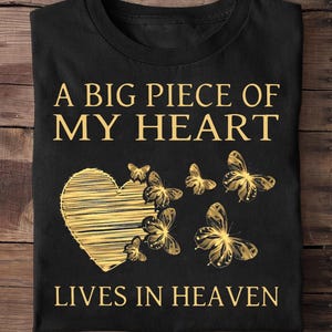 May include: Black t-shirt with gold text and graphics. The text reads "A BIG PIECE OF MY HEART LIVES IN HEAVEN." A gold heart and butterflies are featured in the design. The shirt is folded on a wooden surface.