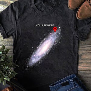 Op de afbeelding: Een antracietgrijs T-shirt met korte mouwen voorzien van een afbeelding van een spiraalstelsel met een rood kaartspeld-icoon dat naar een specifieke locatie wijst. De tekst "YOU ARE HERE" staat in witte hoofdletters boven de afbeelding.