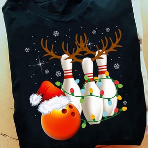 May include: Black t-shirt featuring a Christmas-themed bowling design. The graphic includes bowling pins with reindeer antlers, a bowling ball wearing a Santa hat, and string lights. Snowflakes and stars complete the festive look.