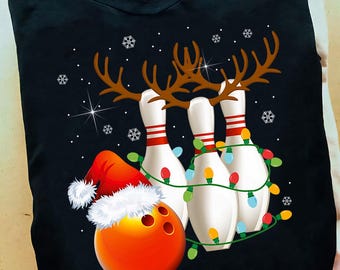 May include: Black t-shirt with a Christmas bowling design. The graphic shows bowling pins with reindeer antlers, a bowling ball in a Santa hat, and fairy lights. Snowflakes and stars complete the festive scene.