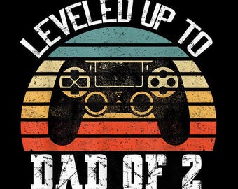 May include: A retro-style graphic with a video game controller and the text "LEVELED UP TO DAD OF 2". The graphic has a sunset background with a red, orange, and yellow color scheme.
