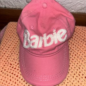 Puede incluir: Gorra de béisbol rosa con la palabra "Barbie" bordada en blanco en la parte delantera. La gorra tiene una visera curva y un botón en la parte superior. Descansa sobre una manta de ganchillo color melocotón.