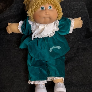 Peut inclure: Une poupée Cabbage Patch Kids vintage vêtue d'une tenue en velours vert d'eau avec une bordure en dentelle blanche et des baskets blanches. La poupée a des cheveux blonds en fil, des yeux bleus et un petit nœud violet. Le nom de la poupée est brodé sur la tenue.