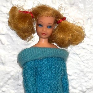 Könnte beinhalten: Eine Vintage-Puppe mit blonden Zöpfen, die mit roten Bändern gebunden sind. Die Puppe hat blaue Augen und trägt einen türkisfarbenen, schulterfreien Pullover. Ein Sammlerstück.