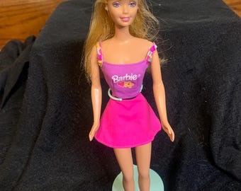 May include: A Barbie doll wearing a pink outfit. The doll has blonde hair and is wearing a pink top with the word "Barbie" on it, a pink skirt, and pink shoes. The doll is standing on a green stand.