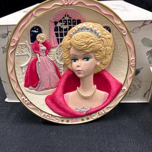 May include: A decorative plate featuring a blonde woman with a tiara and pearl necklace, wearing a red cape. A second figure in a pink dress is in the background. The plate has a pink and gold border.