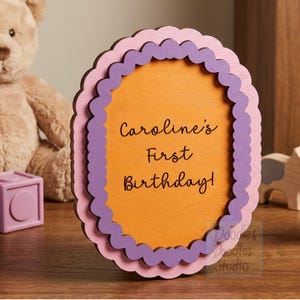 Peut inclure: Un panneau ovale en bois avec un bord festonn&eacute; en couches rose clair et violet. Le centre est orange avec le texte noir "Caroline's First Birthday!" imprim&eacute; en police cursive. Le panneau est pos&eacute; debout sur une surface plane.