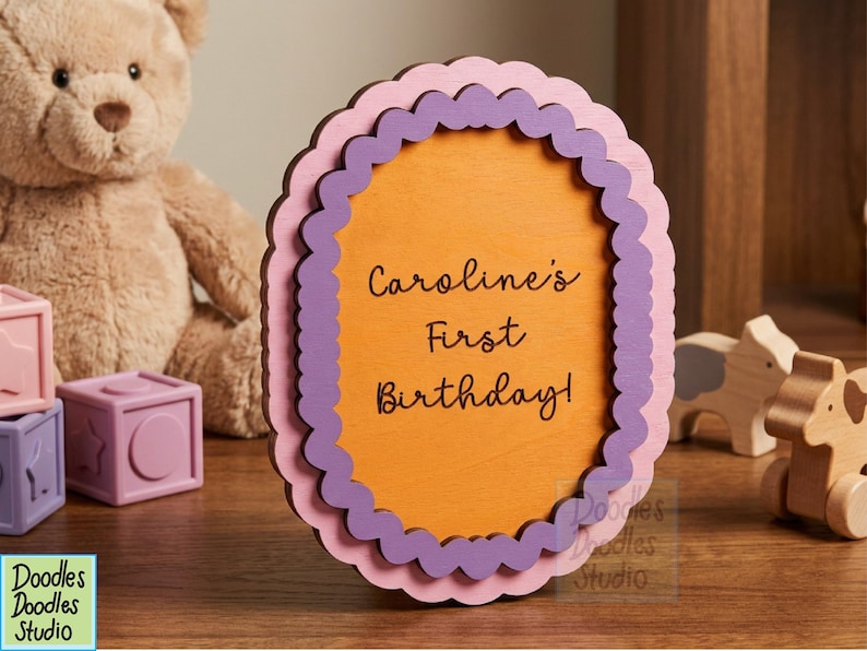 Peut inclure: Un panneau ovale en bois avec un bord festonn&eacute; en couches rose clair et violet. Le centre est orange avec le texte noir "Caroline's First Birthday!" imprim&eacute; en police cursive. Le panneau est pos&eacute; debout sur une surface plane.