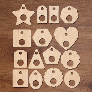 Puede incluir: Un conjunto de etiquetas de madera en varias formas, incluyendo una estrella, un rectángulo y un corazón, cada una con un agujero circular y un agujero más pequeño en la parte superior. Las etiquetas están dispuestas sobre una superficie de madera marrón.