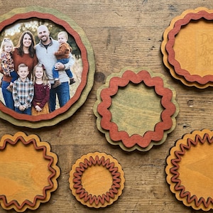 Peut inclure: Une collection de cadres circulaires en bois aux bords festonn&eacute;s dans des tons marron, beige et vert. Un cadre contient une photographie d'une famille de cinq personnes. Un petit panneau carr&eacute; bleu avec le texte Doodles Doodles Studio est visible.